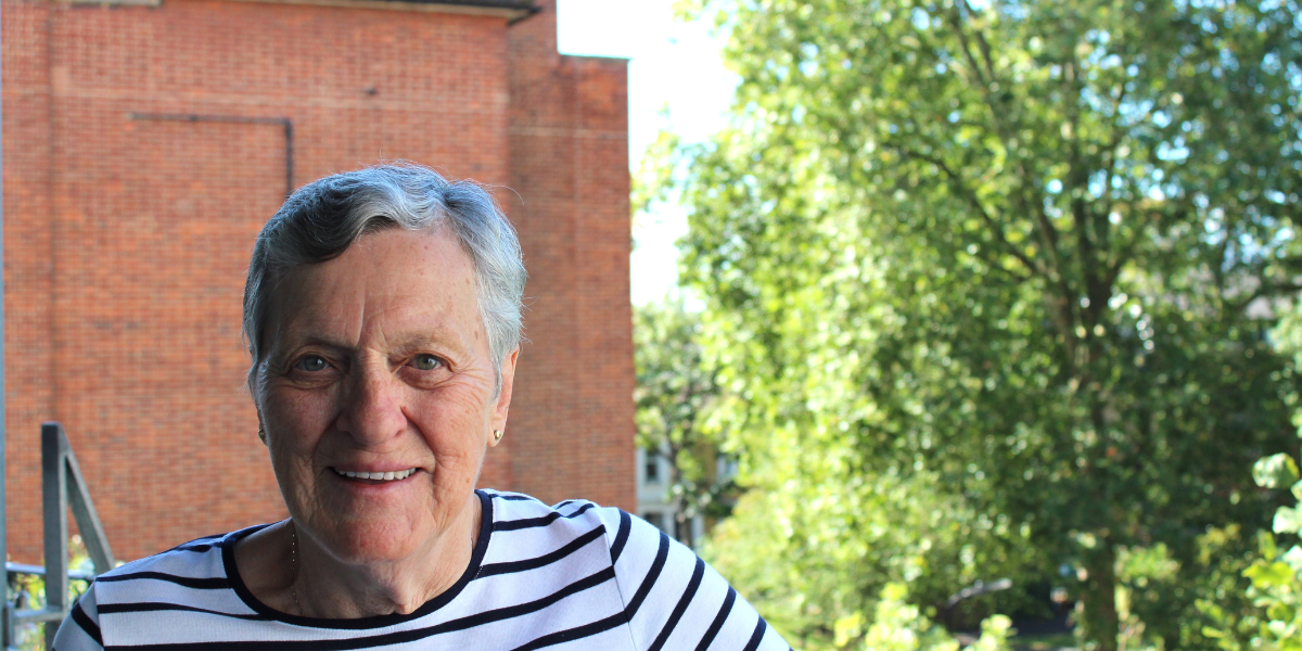 Muriel smiles on her balcony, leafy green trees are in the background. 