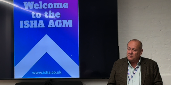 Bob Heapy, is speaking, and standing in front of a board that has a sign which reads Welcome to the ISHA AGM