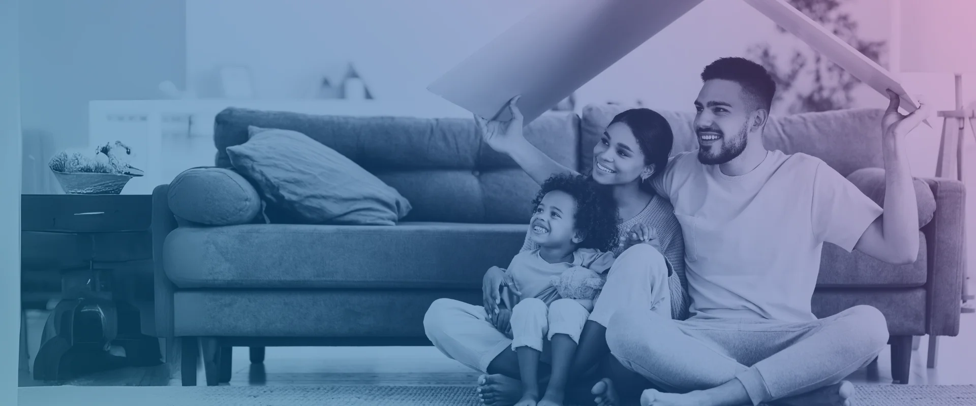 A smiling family sitting in front of a sofa, holding a cardboard above their head in the shape of a roof.